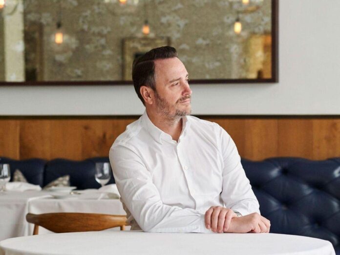 Chef Jason Atherton em um restaurante, vestindo uma camisa branca com um ambiente elegantemente decorado ao fundo.