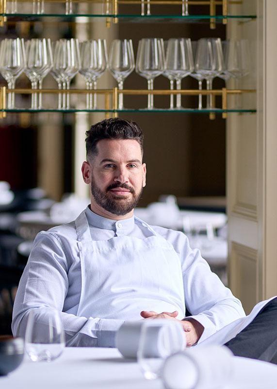 Chef Vincent Crepel posando em um ambiente elegante de restaurante, com mesas organizadas ao fundo