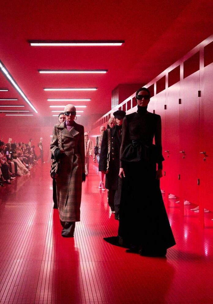 Desfile de moda com fundo vermelho, armários visíveis e modelos em destaque.