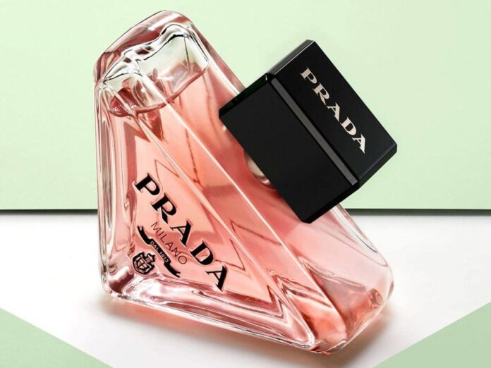 Frasco de perfume rosa com detalhes em vidro e logotipo da Prada, em um fundo suave com cores verde claro e branco.