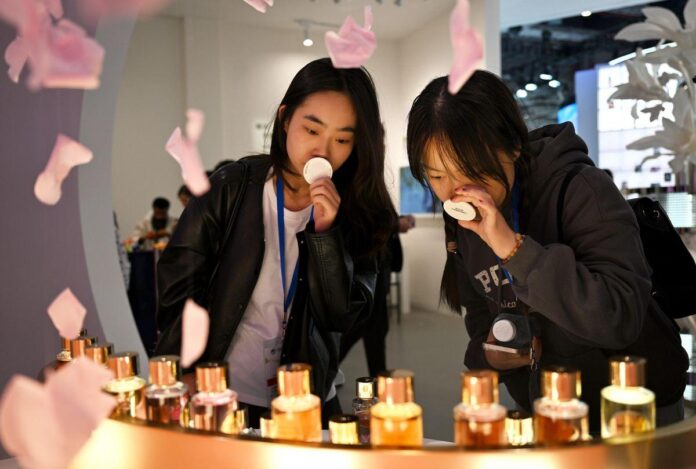 Frascos de perfume em uma mesa com pétalas de rosa caindo ao redor e mulheres sniffing amostras de fragrâncias.