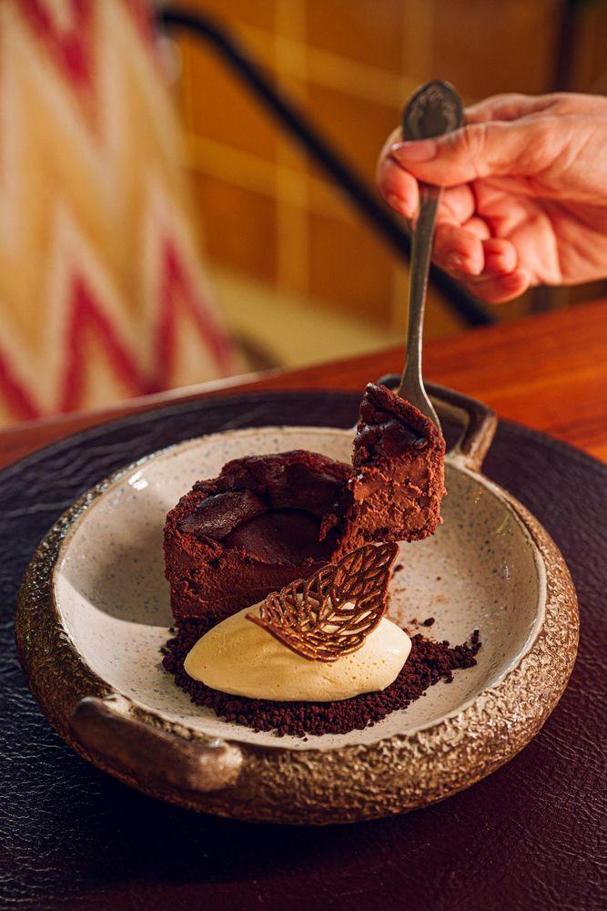 Imagem do doce de chocolate, com creme claro e decoração de chocolate em forma de folha, em um prato raso. O fundo desfocado retrata um ambiente acolhedor de restaurante.