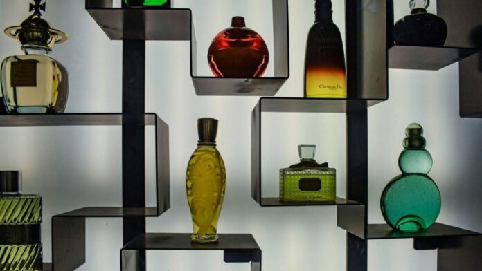 Imagem frontal de frascos de perfume coloridos em prateleiras geométricas, com fundo branco iluminado.