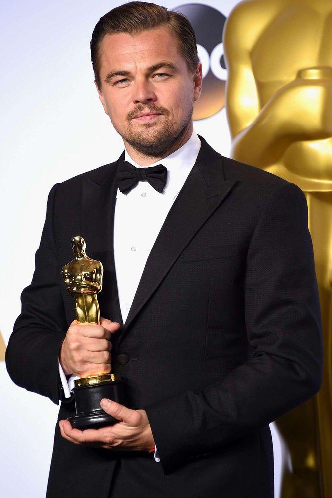 Leonardo DiCaprio usando um tônico preto e gravata borboleta, segurando a estatueta do Oscar, com um fundo suave e desfocado.
