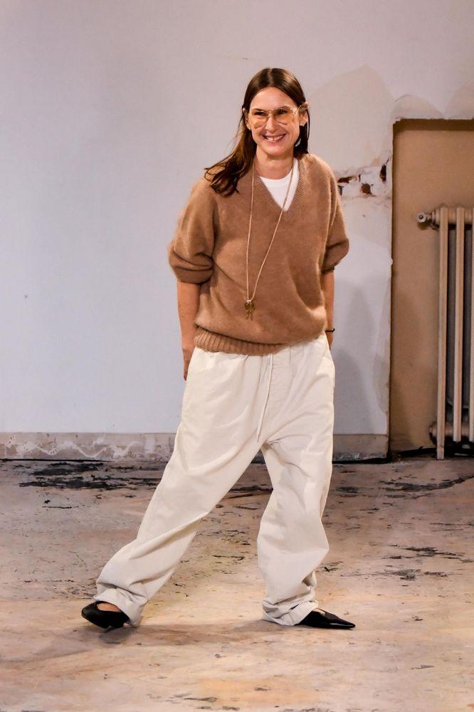 Louise Trotter sorrindo, vestindo um suéter marrom e calças largas brancas, com óculos e cabelo solto, com fundo de parede clara e um radiador visível.