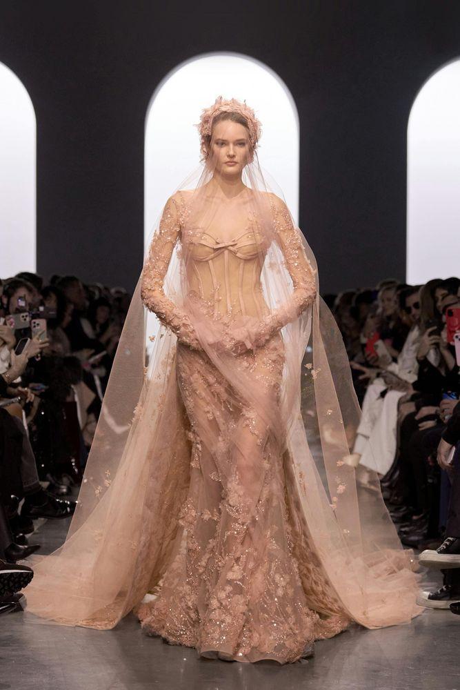 Modelo usando um vestido de noiva em tons de rosa claro com detalhes florais, em um fundo escuro que destaca a peça.