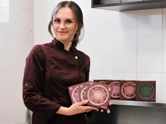 Mulher com cabelo solto e óculos usando jaleco marrom em uma cozinha profissional, com caixas de chocolate coloridas ao fundo.