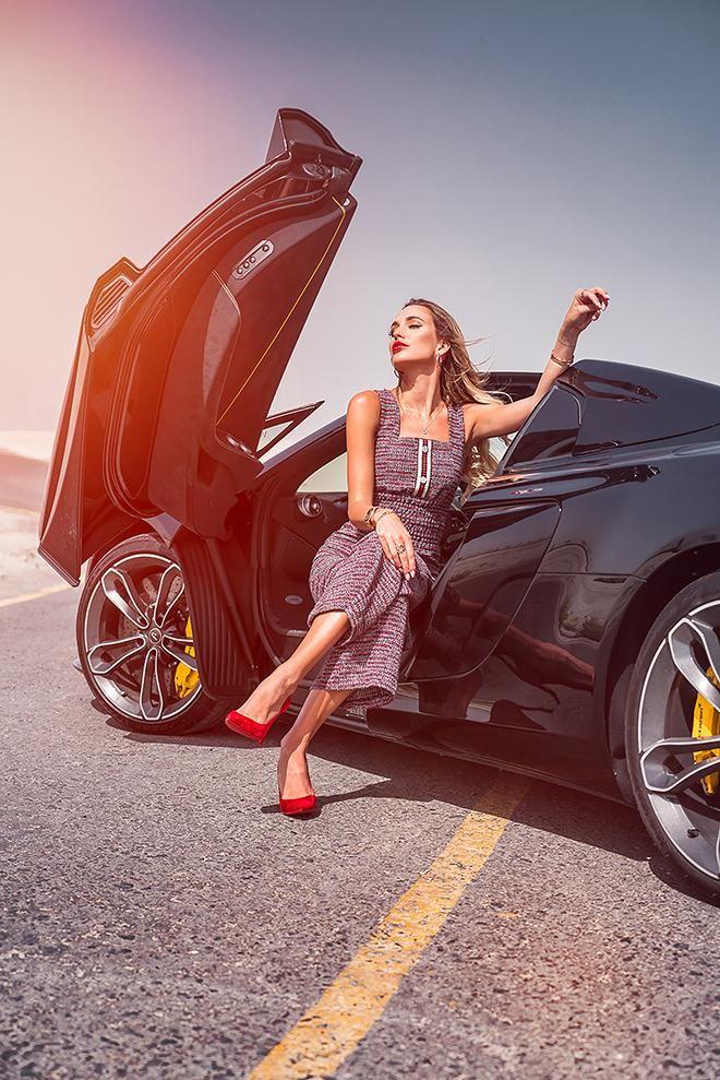 Mulher posando ao lado de um supercarro preto em uma estrada clara com o céu azul.