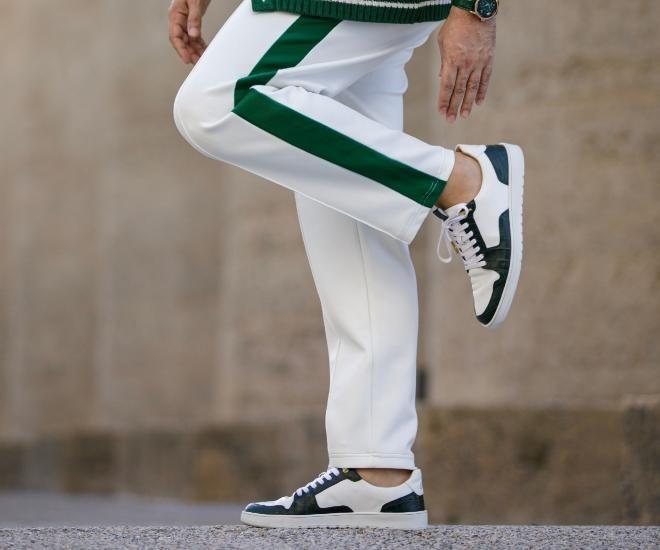 Tênis branco e verde sendo usado por uma pessoa em movimento, com uma calça branca e detalhes em verde, em um fundo de pedra com textura clara.