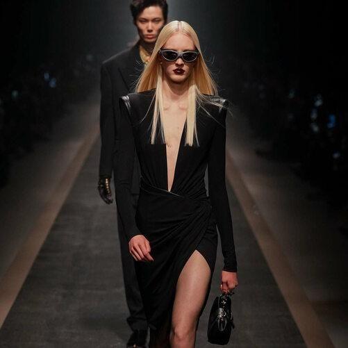 Uma modelo usando um vestido preto com decote profundo e fenda na perna, caminhando na passarela sob iluminação dramática.