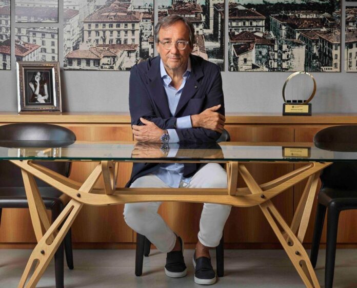 Homem com cabelos grisalhos, usando óculos, jaqueta escura e camisa clara, sentado em uma mesa de vidro ao lado de uma estatueta e uma foto emoldurada, em um fundo neutro e minimalista.